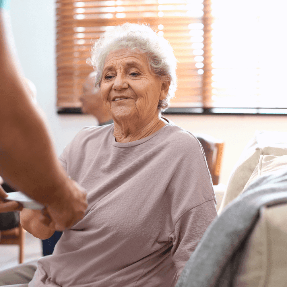An elderly woman with short white hair smiles while sitting on a couch as a person hands her something. - Home Instead