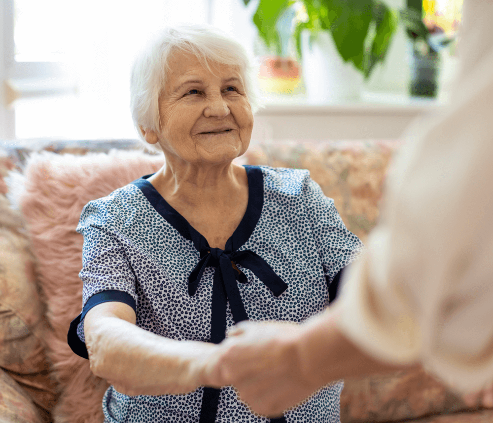 Elderly woman with short white hair smiling and holding hands with another person in a cozy room. - Home Instead