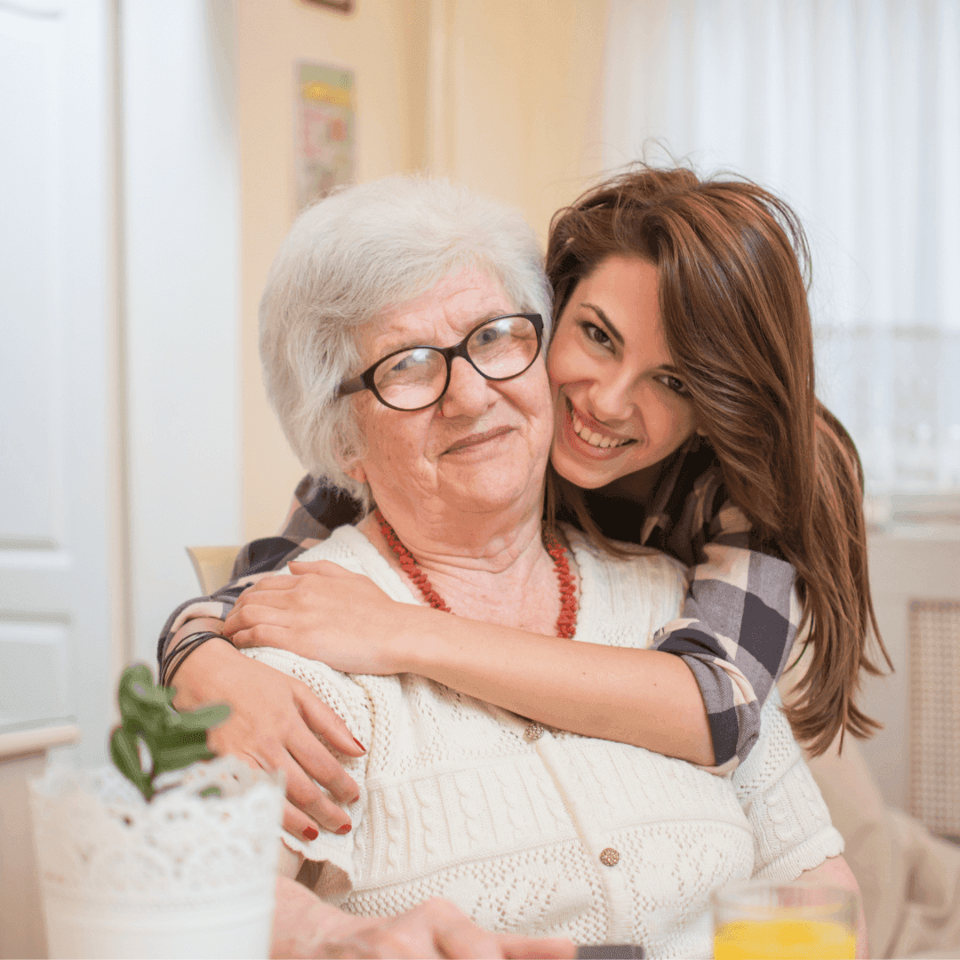 An elderly woman with a younger woman smiling and hugging her from behind in a cozy, bright room. - Home Instead