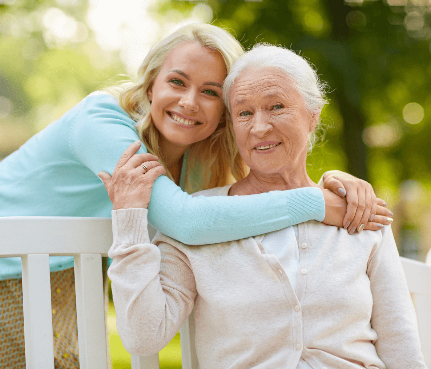 Young woman hugging elderly woman from behind on a park bench, both smiling. Background is green and sunny. - Home Instead