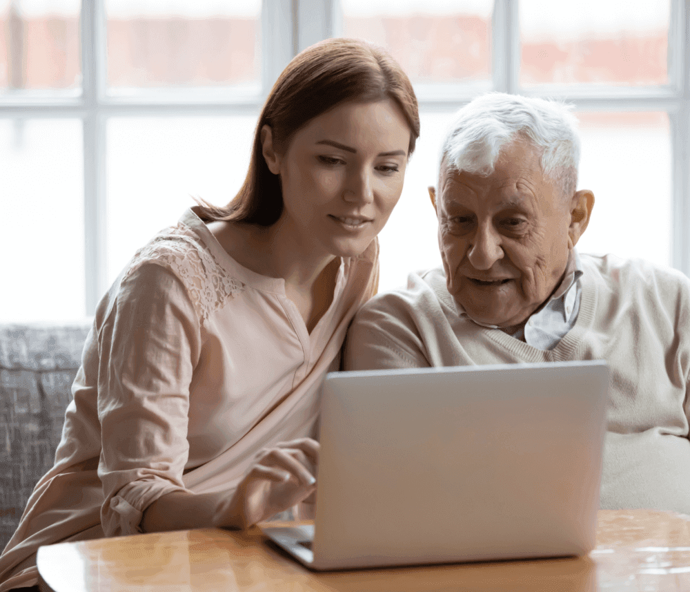 A young woman and an elderly man sit together, smiling and looking at a laptop screen. - Home Instead