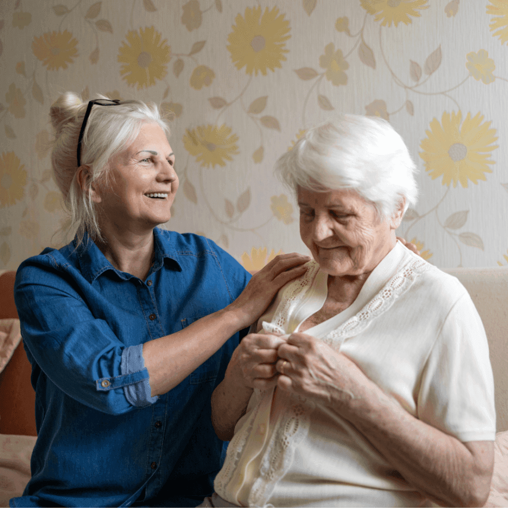A smiling woman comforts an elderly woman seated on a couch. - Home Instead