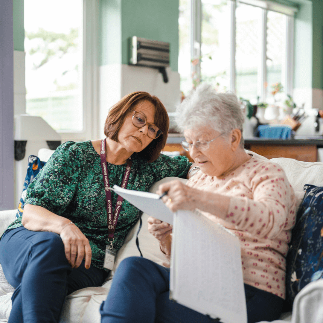 Two elderly women sitting on a couch, reading a document together, in a brightly lit room with plants and windows. - Home Instead