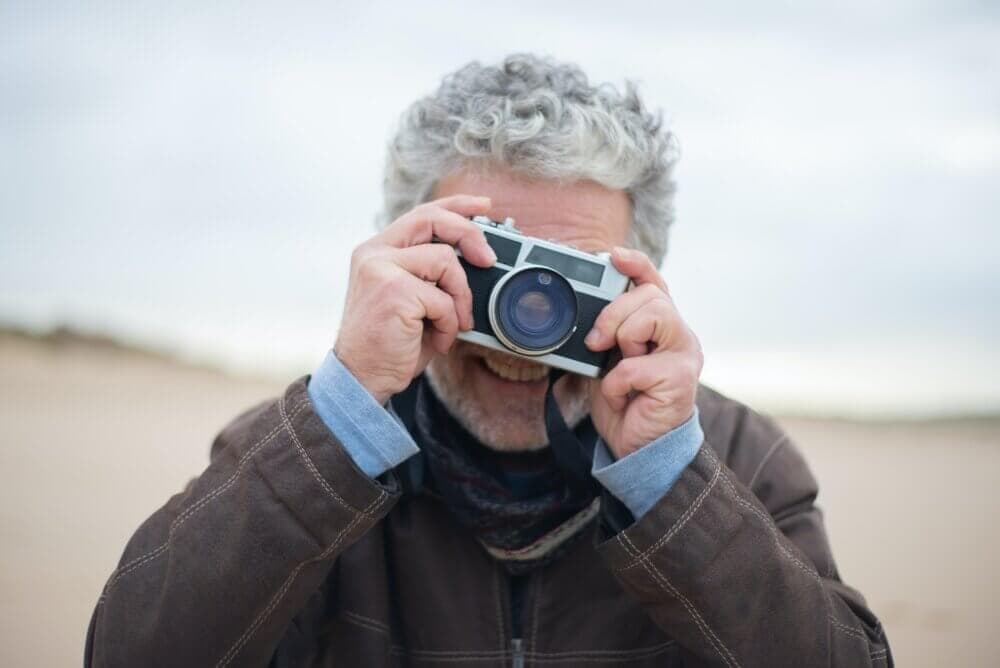 A person with curly gray hair takes a photo with a vintage camera outdoors, wearing a brown jacket and blue shirt. - Home Instead