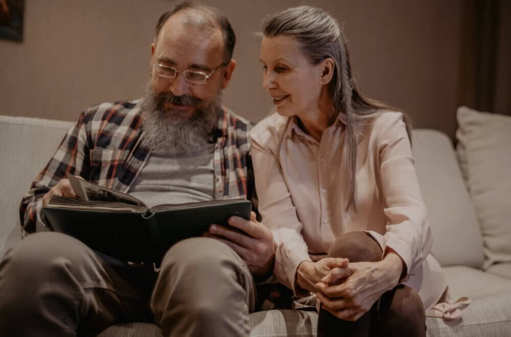 An elderly couple sitting on a couch, looking at a photo album together, smiling and reminiscing. - Home Instead