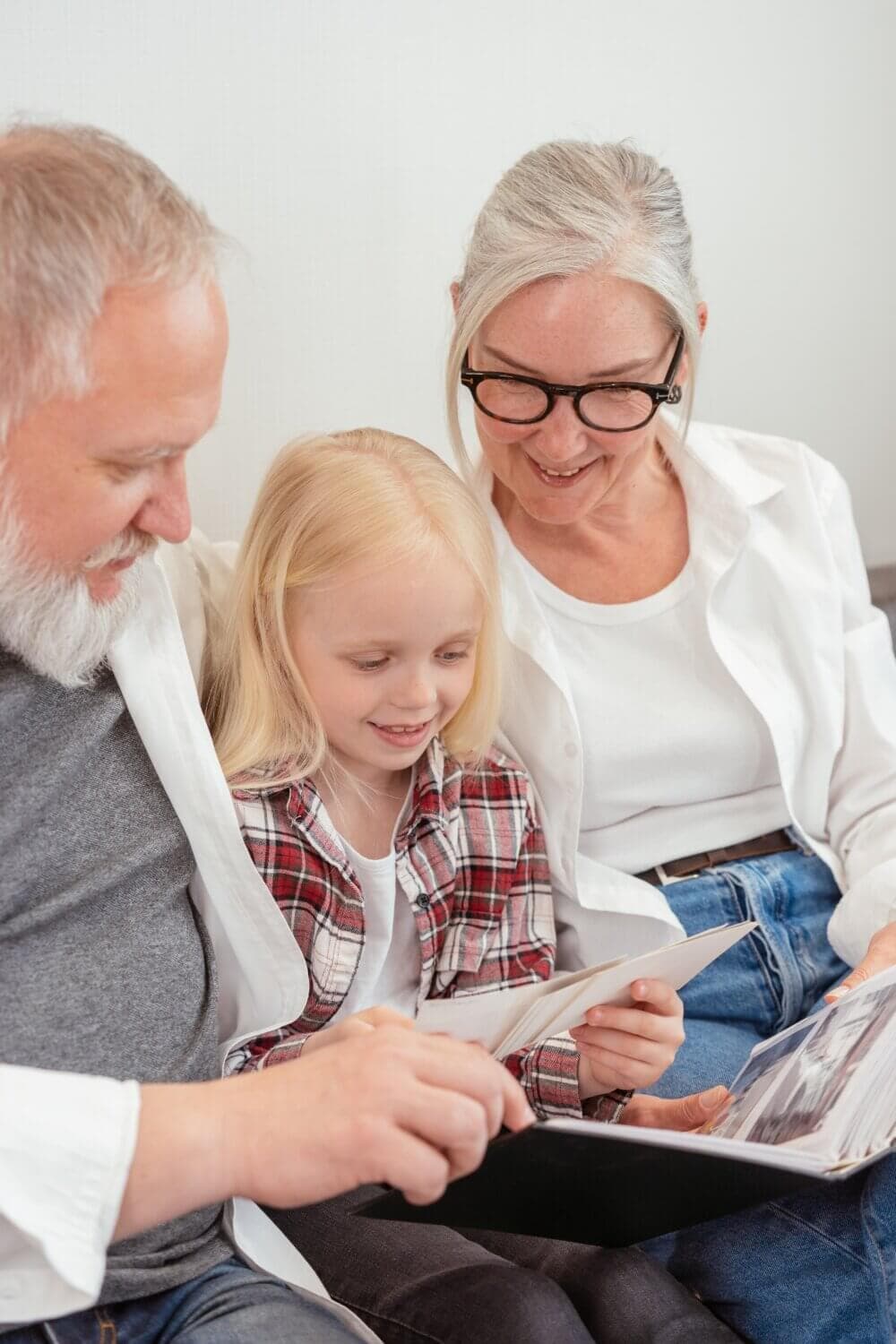 Elderly couple and young girl sitting together, looking at an open photo album, smiling and reminiscing. - Home Instead