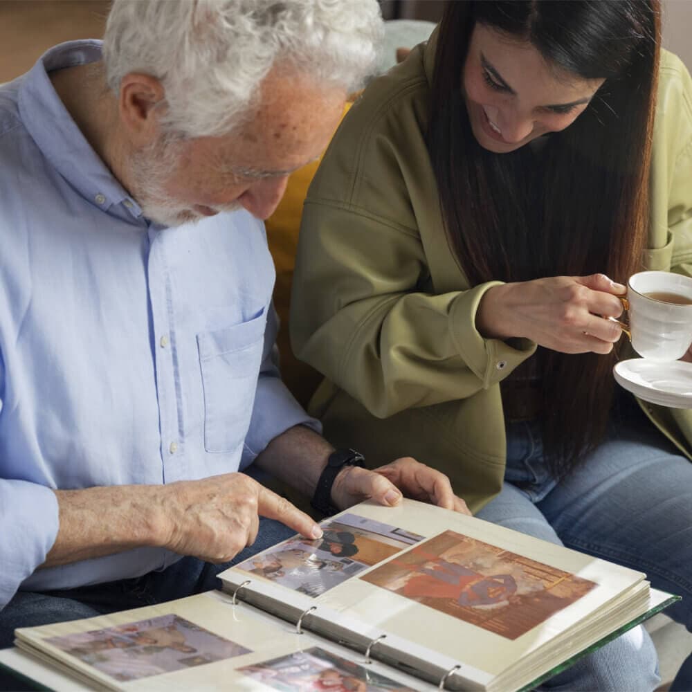 An elderly man and a young woman sit together, smiling and looking at a photo album while having tea. - Home Instead