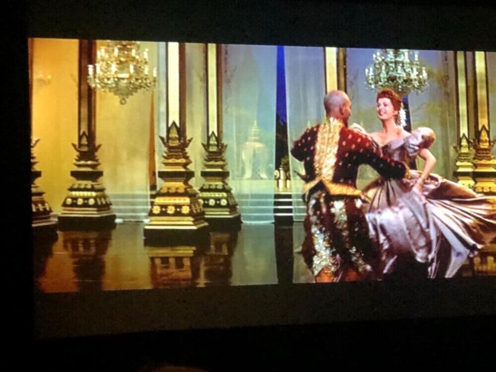 Man in ornate attire dances with a woman in a ballroom. Opulent chandeliers hang above, and pillars are in the background. - Home Instead