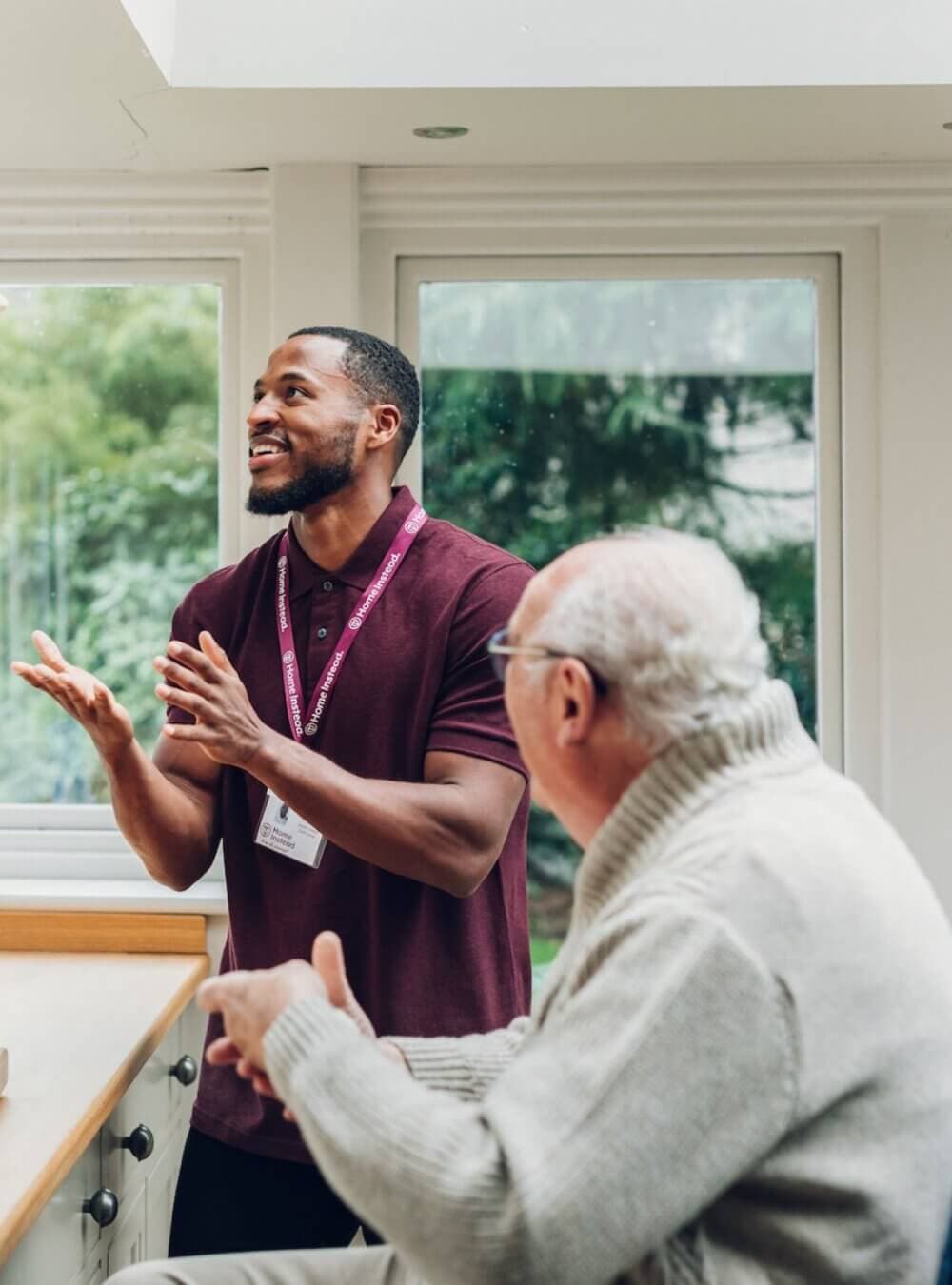 A caretaker in a maroon shirt talks to an elderly man seated in a cozy room with large windows and greenery outside. - Home Instead