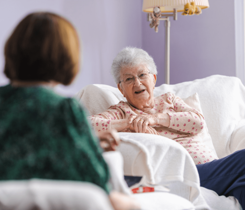 An elderly woman sitting on a white couch, smiling and talking with another person in a green shirt. - Home Instead