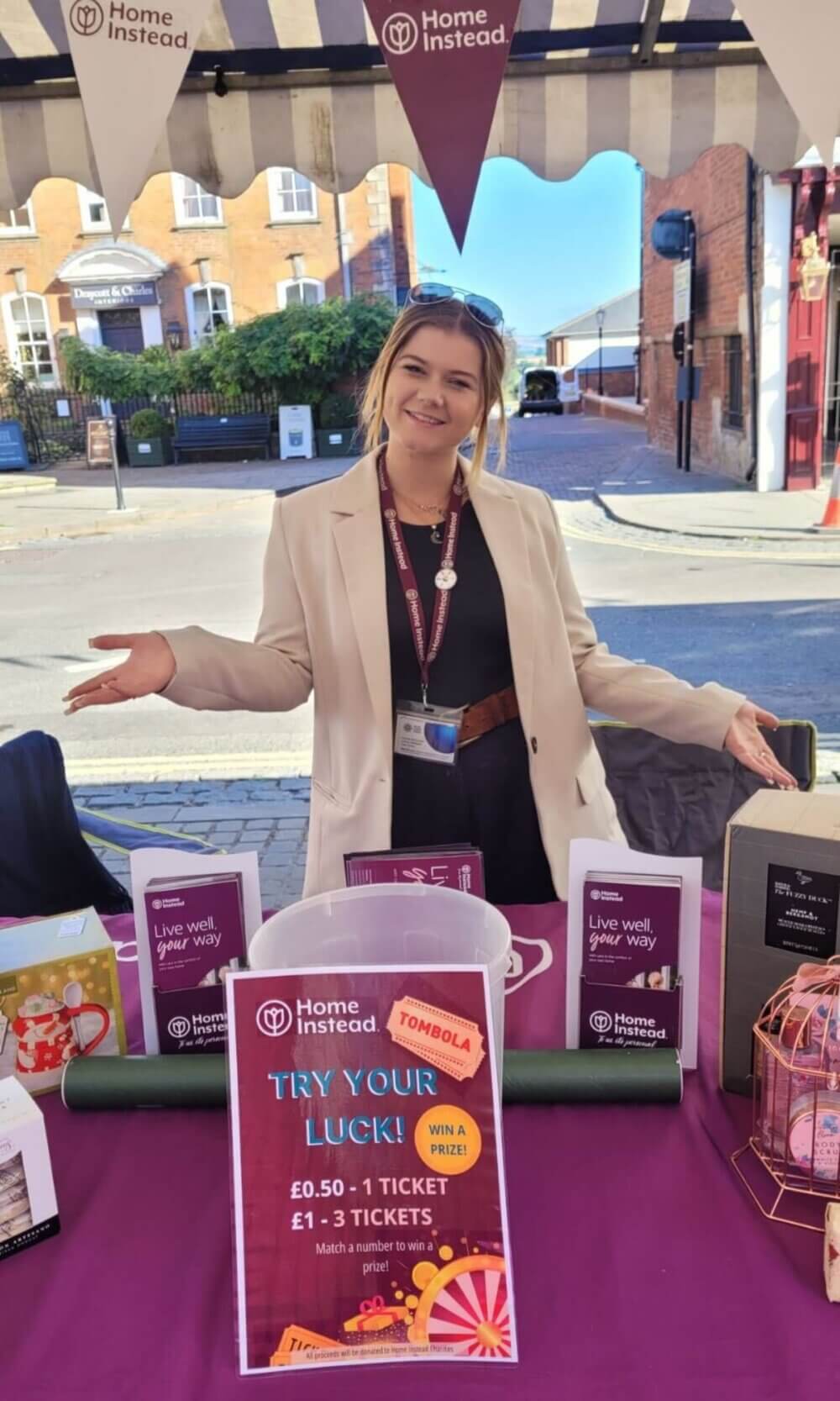 A woman stands at a "Home Instead" booth with a "Try Your Luck" sign, ready to sell tombola tickets. - Home Instead
