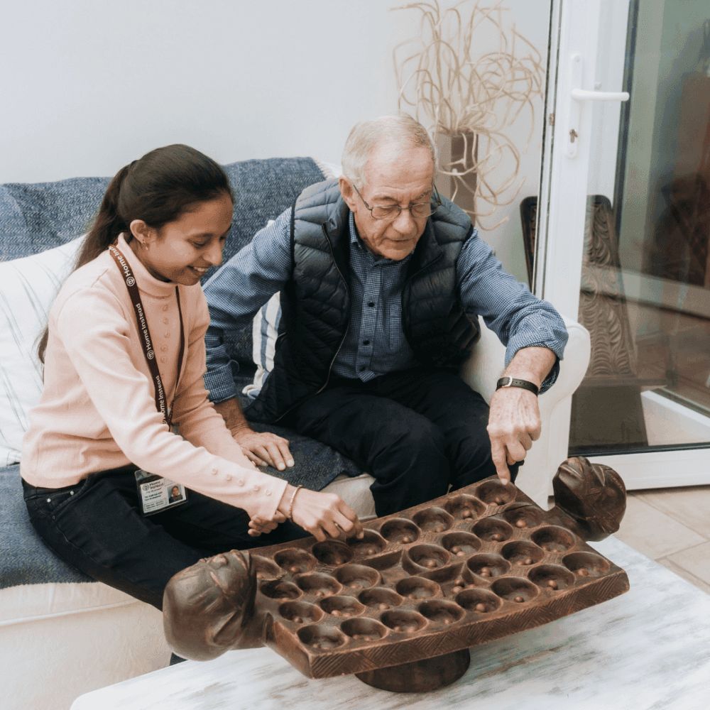 A young girl and an elderly man happily playing a board game together on a table. - Home Instead