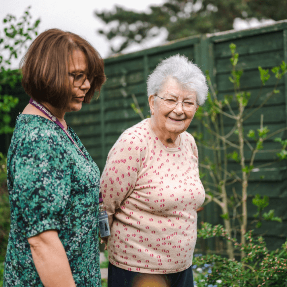 An elderly woman and a younger woman walk together in a garden, surrounded by greenery and wooden fences. - Home Instead