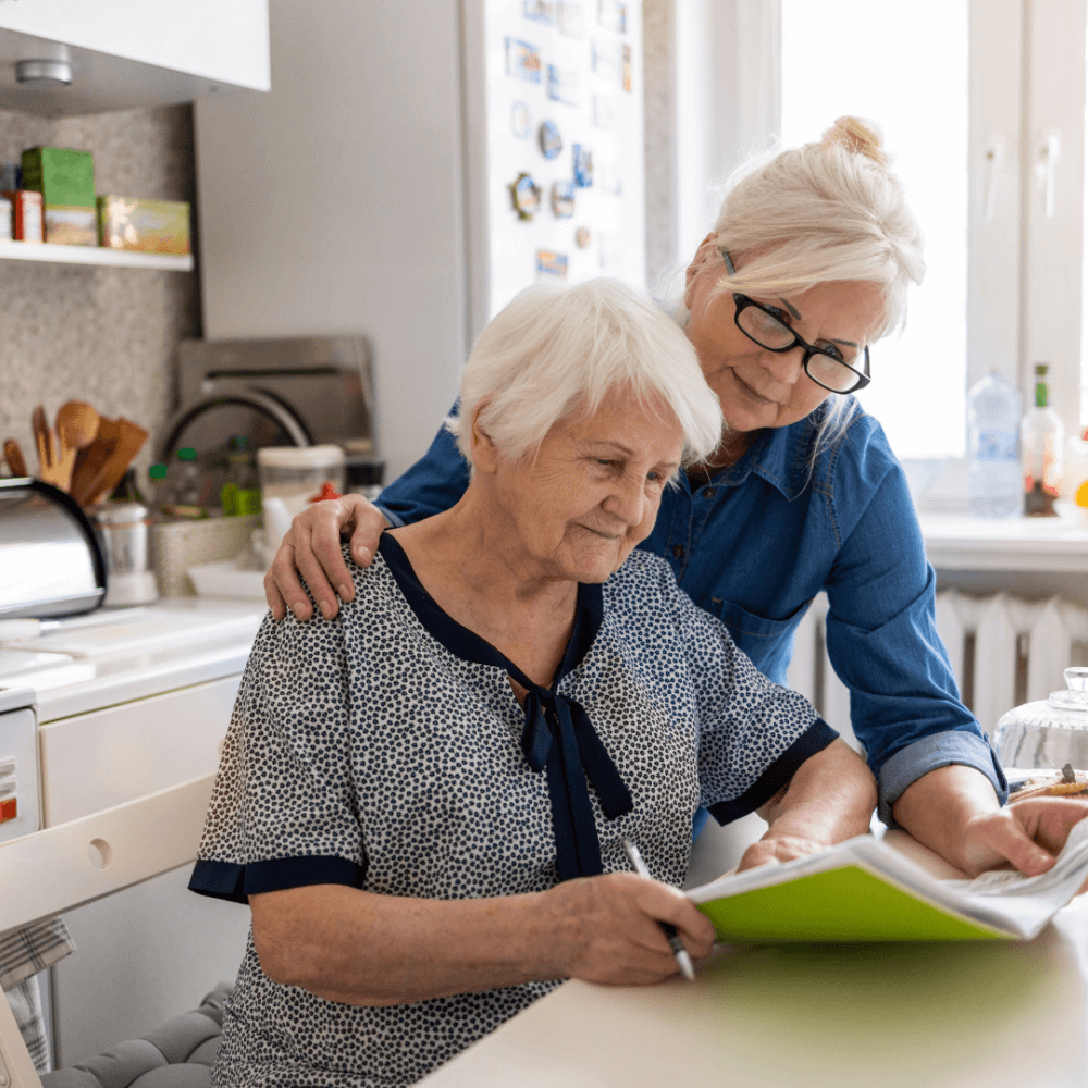 Two elderly women reading a book together in a cozy kitchen. The older woman is seated while the other stands beside her. - Home Instead