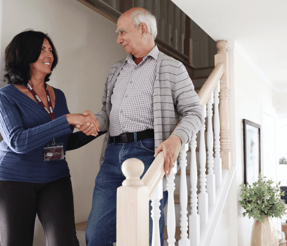 A woman and an older man shake hands near a staircase inside a home, both smiling. The woman wears a name badge. - Home Instead