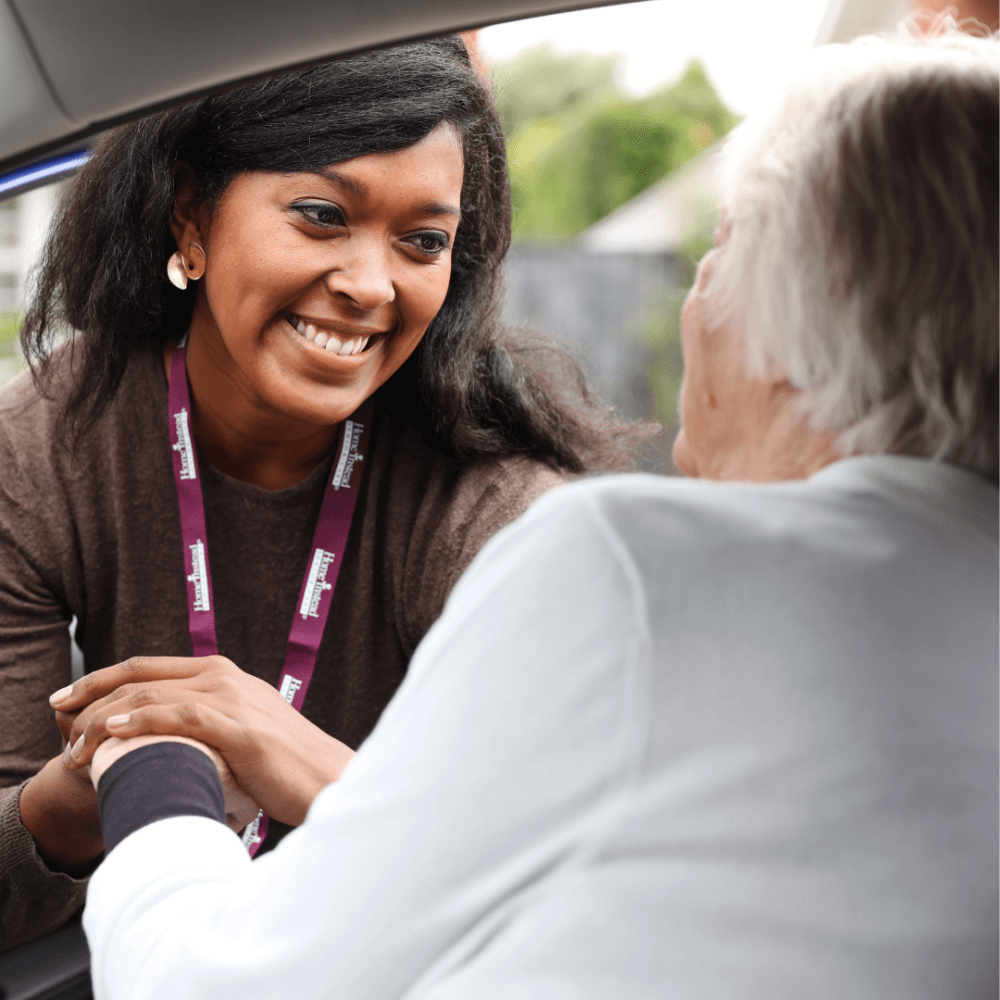 A caregiver with a badge smiles warmly, holding hands with an elderly person who is seated. - Home Instead