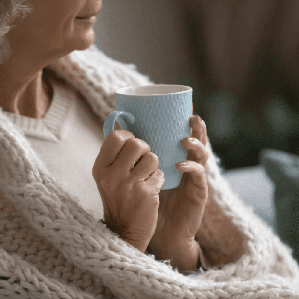 Elderly person wrapped in a cozy blanket, holding a light blue mug with both hands. - Home Instead