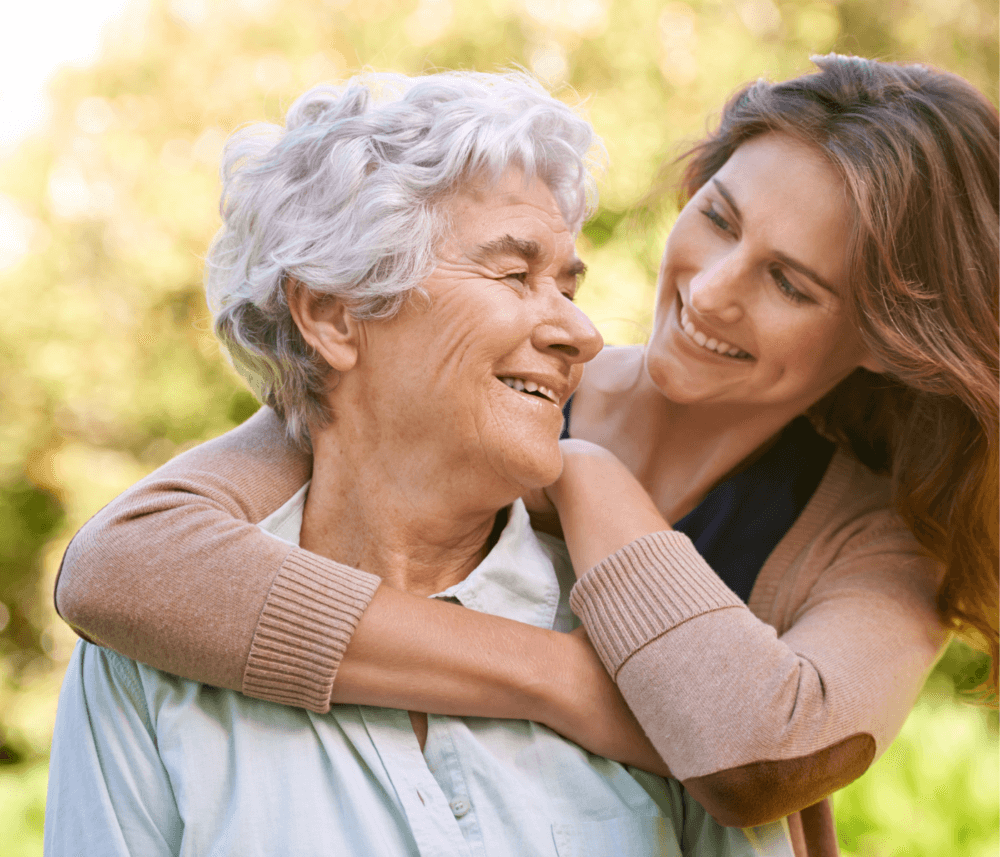 A young woman embraces an elderly woman from behind, both smiling and enjoying an outdoor moment together. - Home Instead