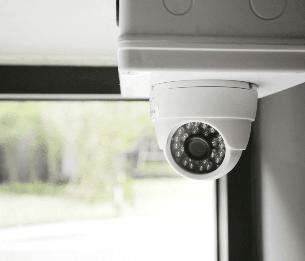 Close-up of a white security camera mounted on a ceiling near a window with a blurred outdoor background. - Home Instead