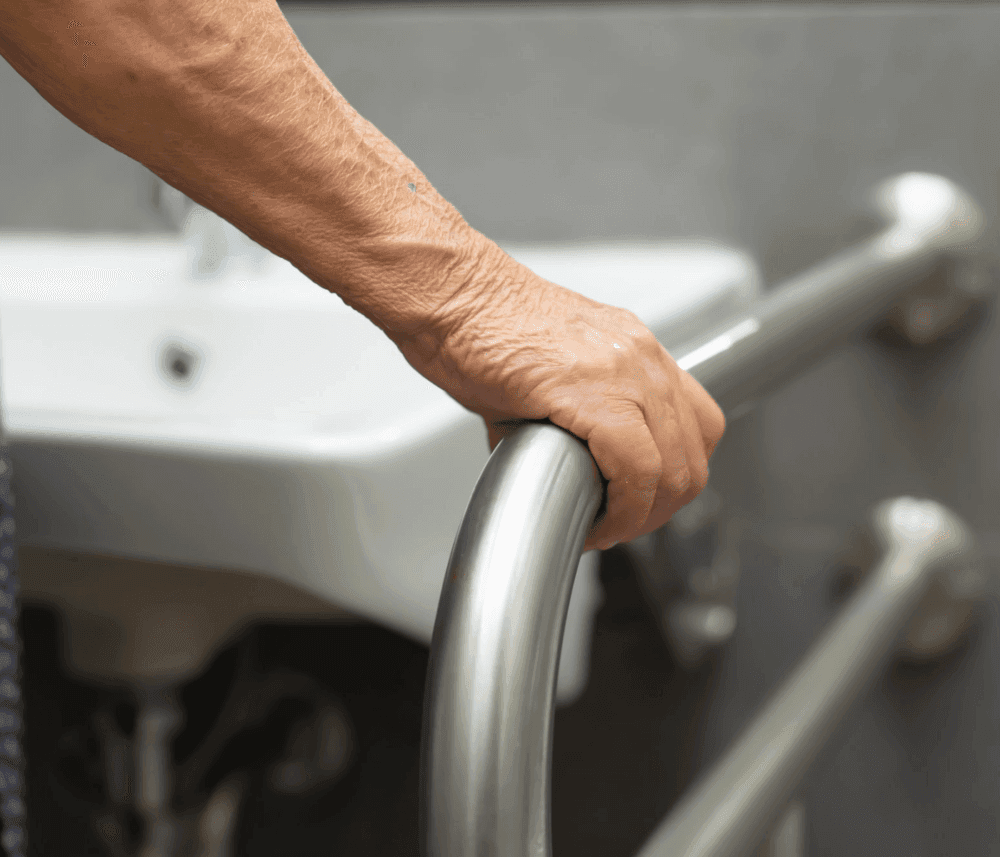 An elderly hand grips a metal handrail in a bathroom, with a sink visible in the background. - Home Instead