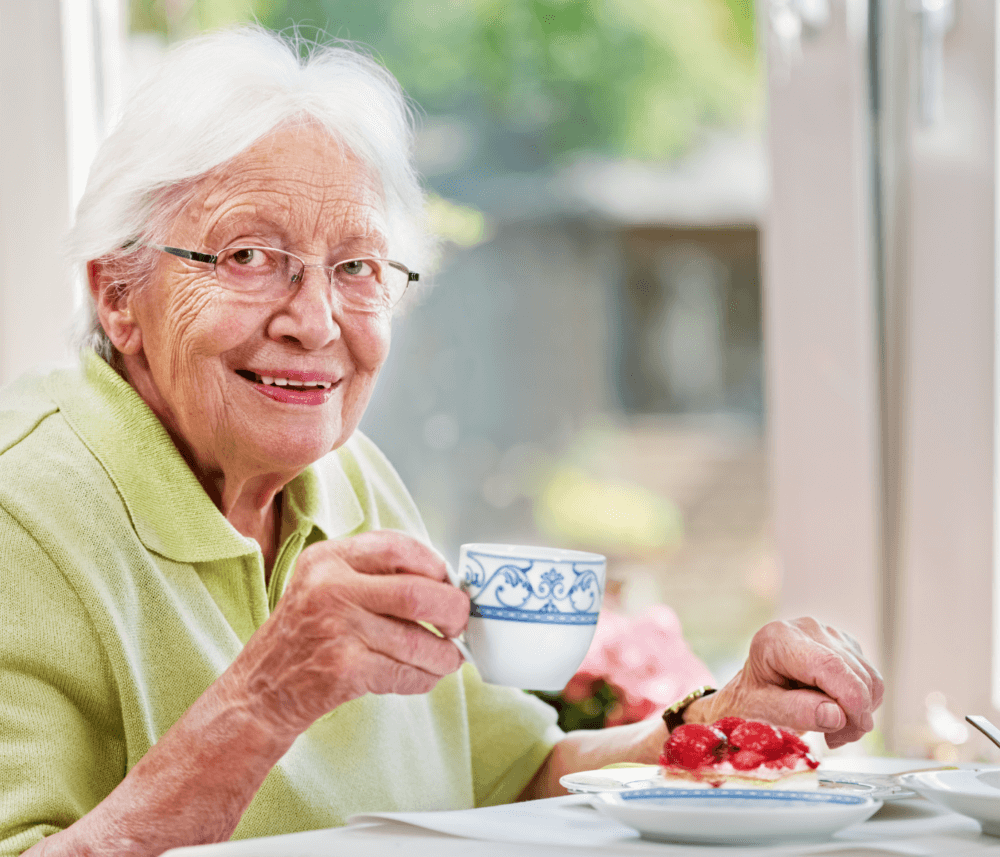Elderly woman with white hair and glasses enjoying a cup of tea and dessert at a table in a bright room. - Home Instead