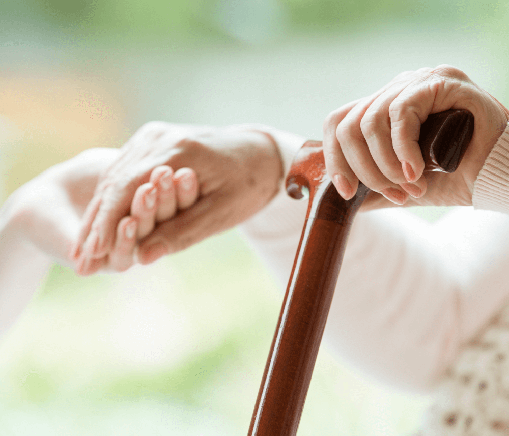 Close-up of elderly hands holding a cane, with one hand resting gently on another for support. - Home Instead