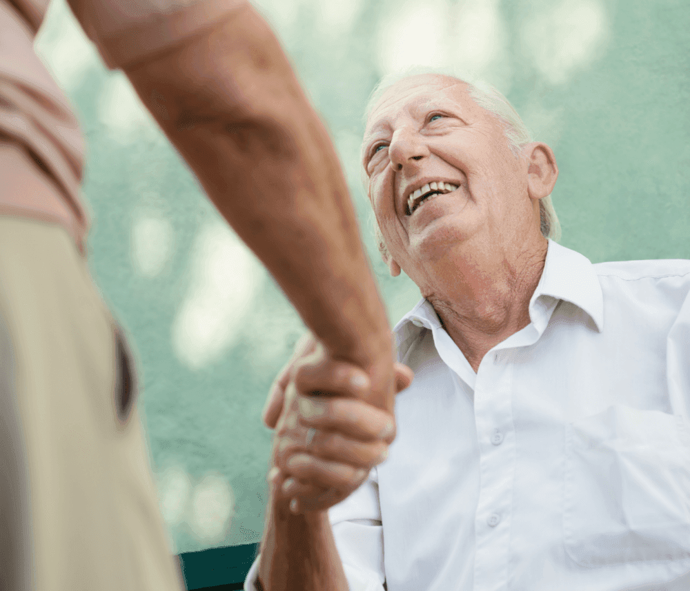 An elderly man wearing a white shirt, seated outdoors, smiles while shaking hands with another person. - Home Instead