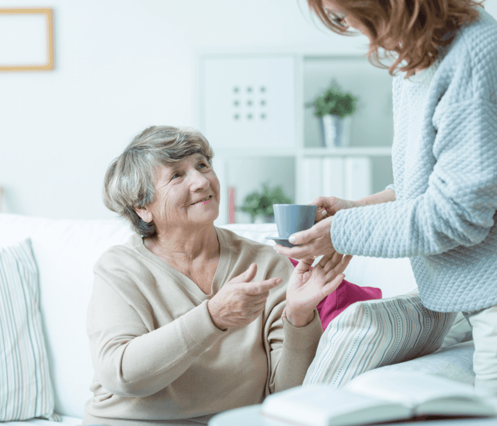 Elderly woman smiling while receiving a cup of tea from a younger person in a cozy living room. - Home Instead
