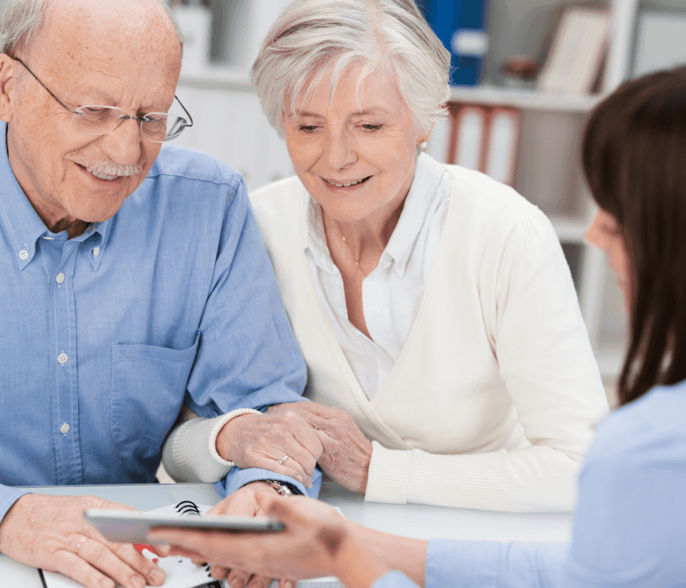 An elderly couple looks at a tablet held by a young woman in a professional setting. - Home Instead