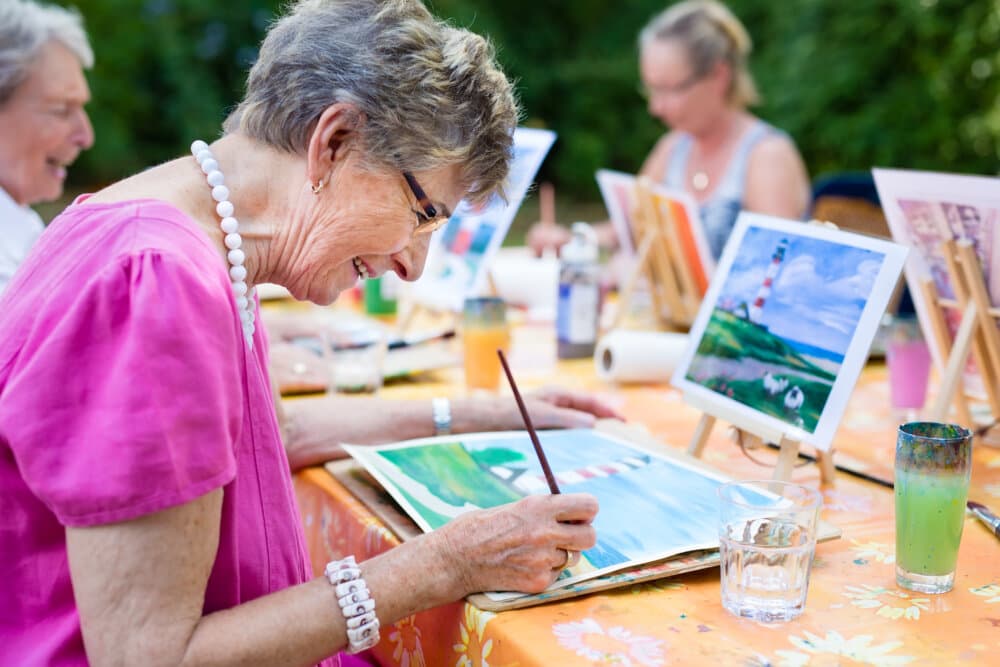 Older woman joyfully painting a landscape on a canvas at an outdoor art class, with others painting in the background. - Home Instead