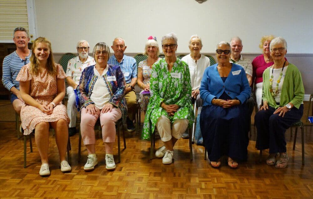 A group of elderly people and two younger individuals are sitting in chairs, smiling at the camera indoors. - Home Instead