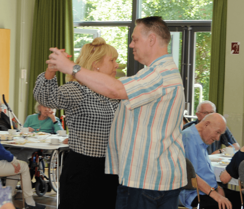 Two people dancing together in a room with seated older adults in the background. - Home Instead