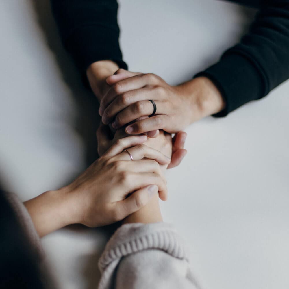 Two pairs of hands gently holding each other on a white surface, showing support and connection. - Home Instead