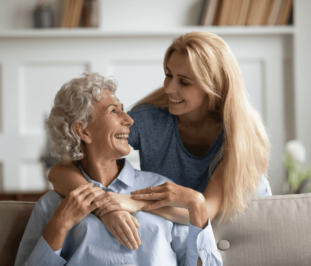 Older woman with gray hair sitting, smiling at younger blonde woman standing behind her, both sharing a loving embrace. - Home Instead