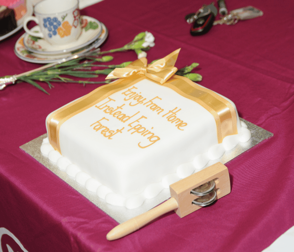 Rectangular cake with white icing, gold ribbon, and text "Enjoy from Home Instead Epping Forest" on a maroon tablecloth. - Home Instead