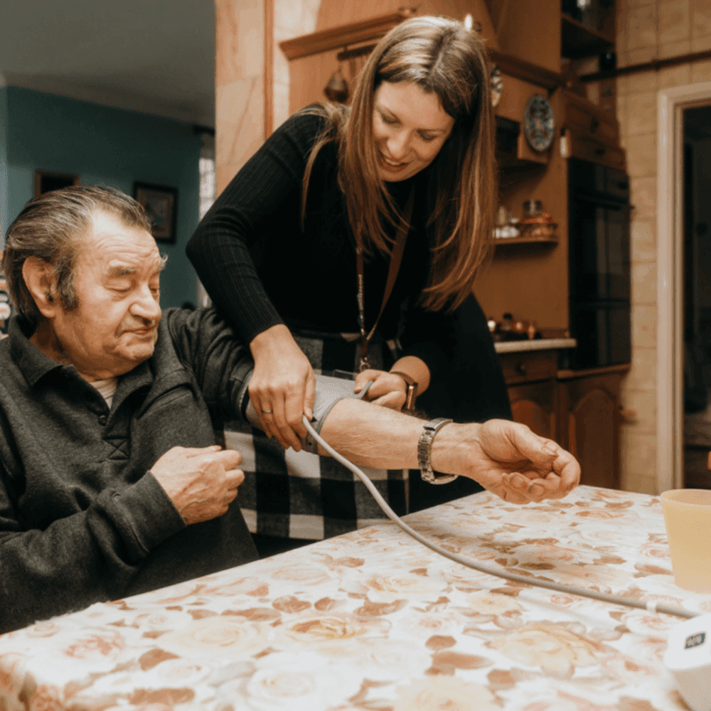 A woman measures an elderly man's blood pressure while they sit at a floral-patterned table in a kitchen. - Home Instead