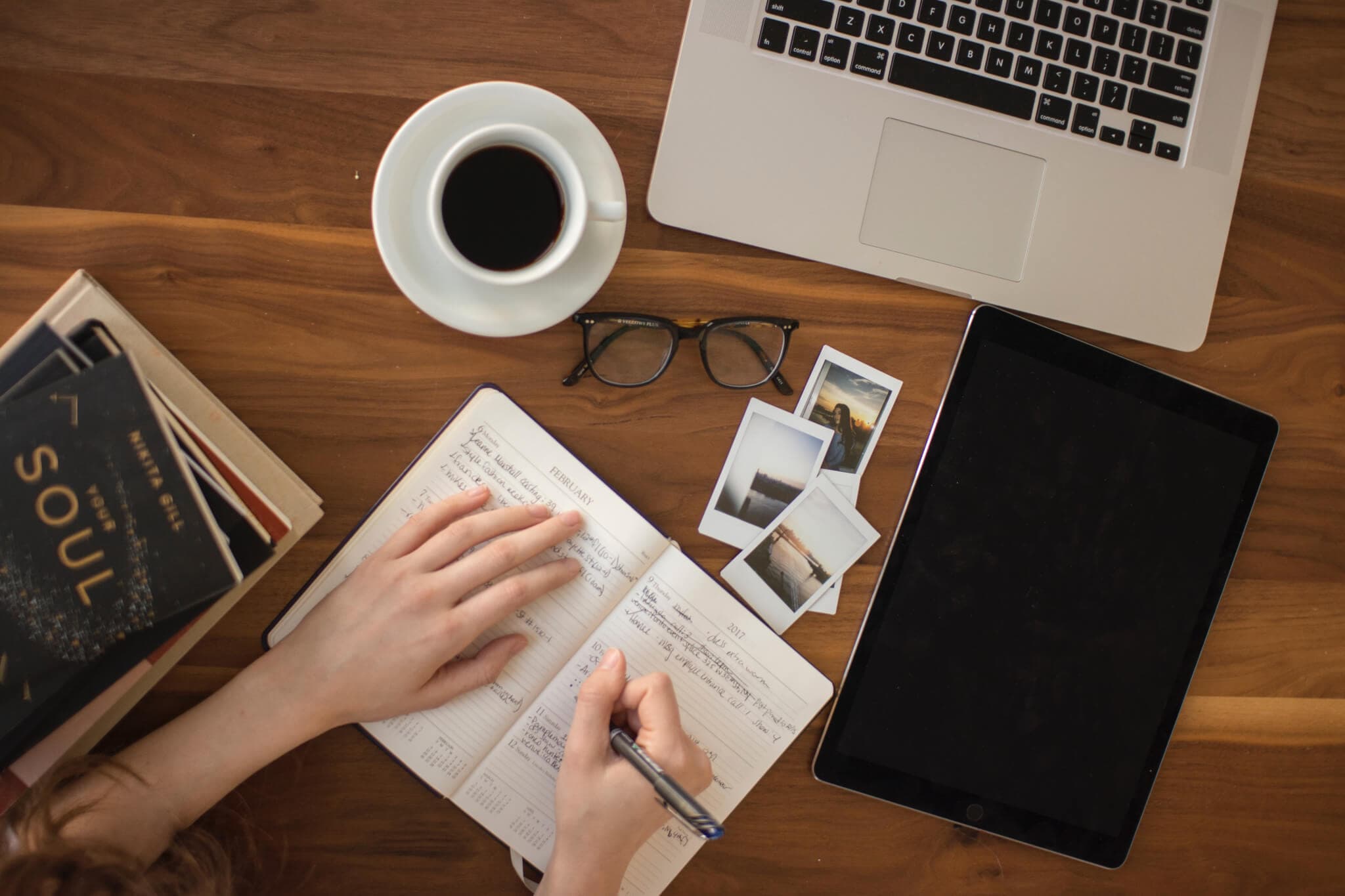 Hands writing in a notebook on a wooden desk with a laptop, tablet, coffee cup, book, glasses, and photos. - Home Instead