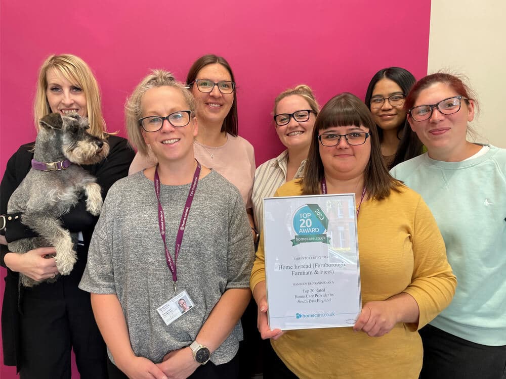 Seven women, one holding a dog, smile at the camera. One woman holds a framed "Top 20 Award" certificate. - Home Instead