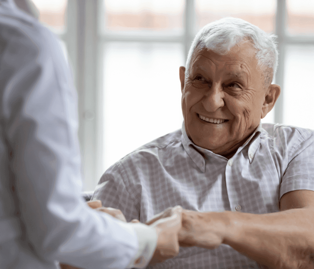 An elderly man smiling at a person wearing a white coat, holding hands in a brightly lit room. - Home Instead