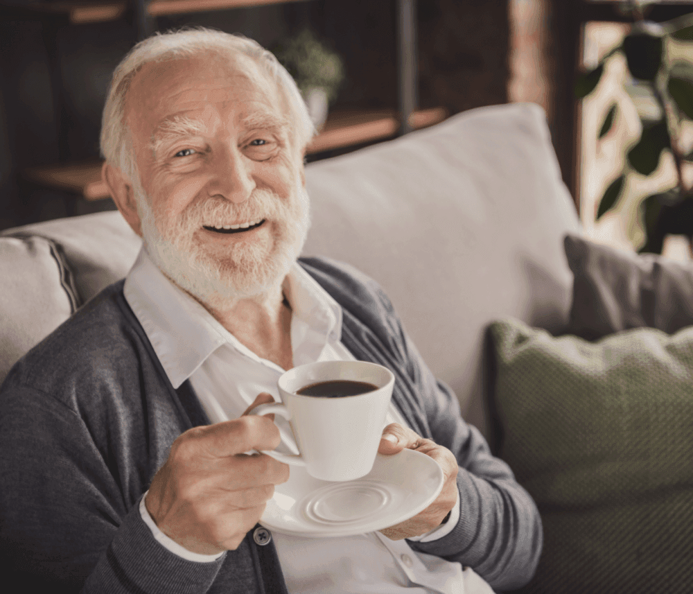 Smiling elderly man with white beard holding a cup of coffee, sitting on a sofa with green cushions behind. - Home Instead