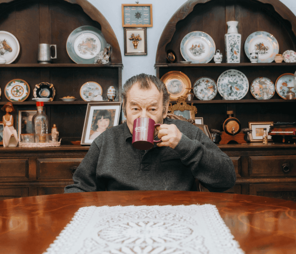 A man sips from a mug at a wooden table with a lace runner, in front of shelves displaying various decorative plates. - Home Instead