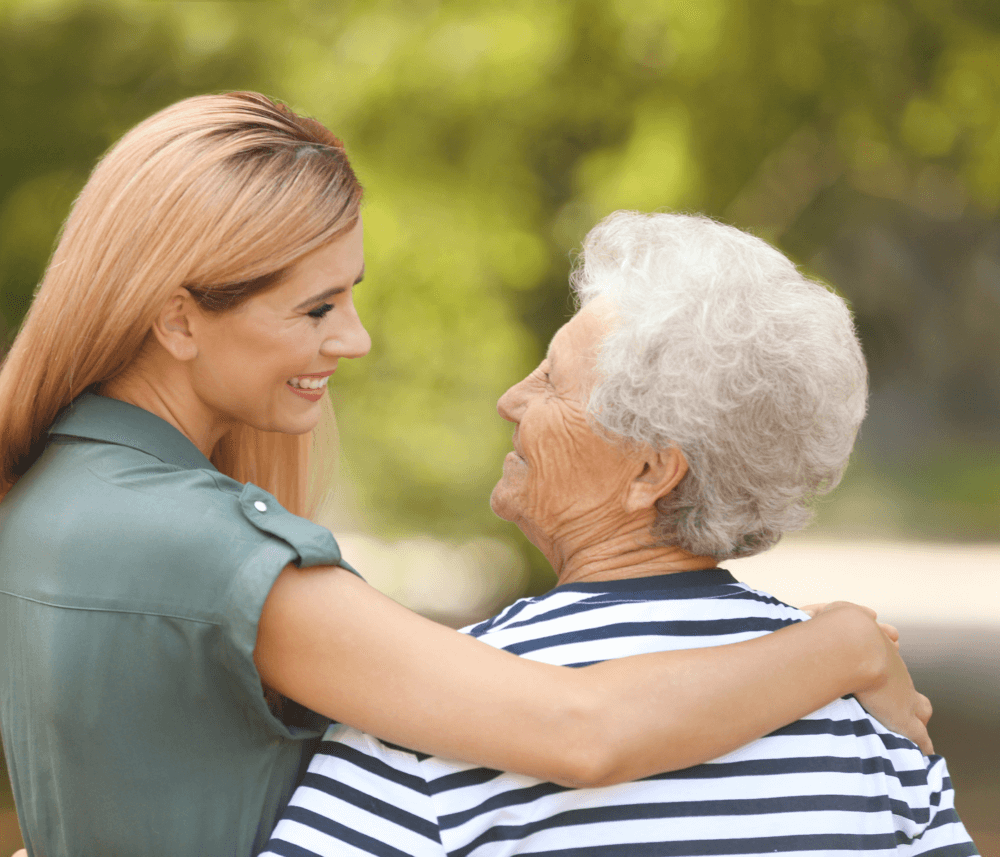 A young woman with blond hair hugs an elderly woman with gray hair outdoors, both smiling at each other. - Home Instead