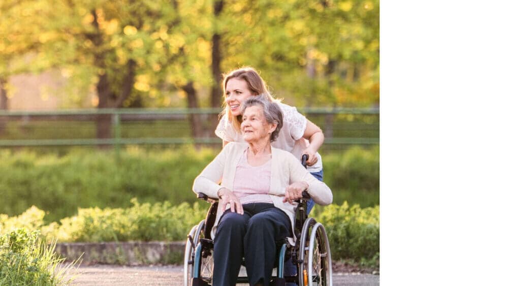A young woman joyfully pushing an elderly woman in a wheelchair through a sunny park. - Home Instead