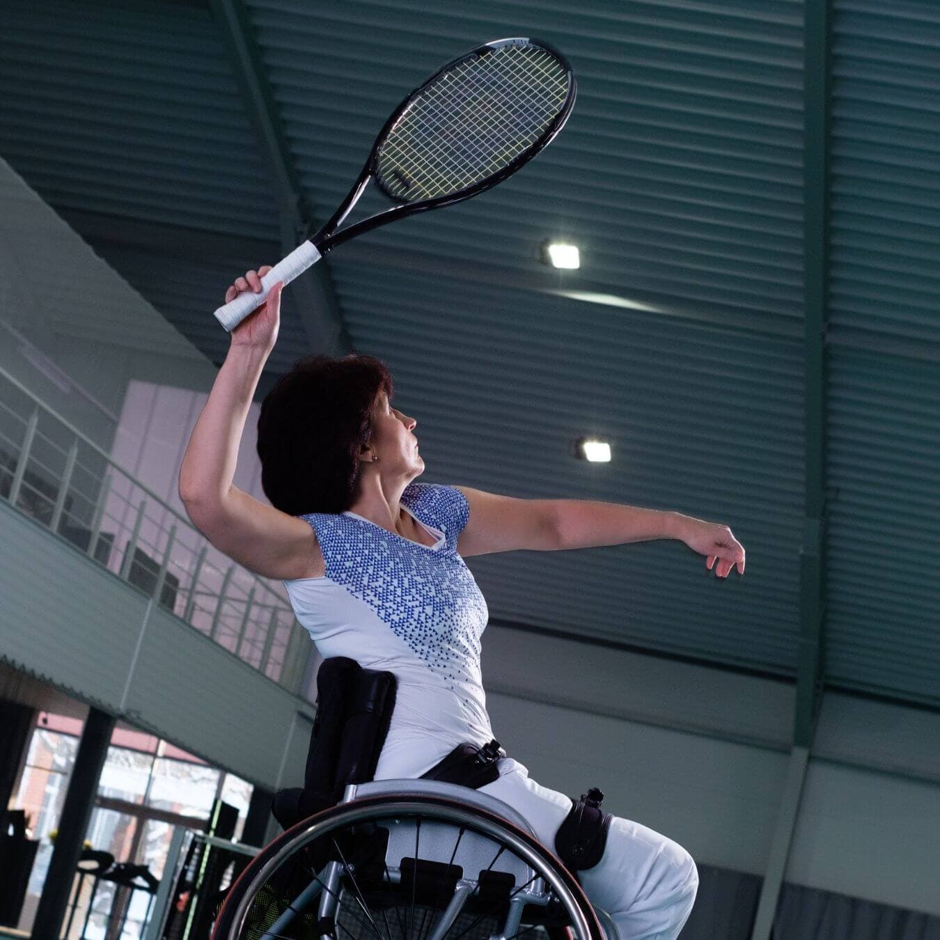 A woman in a wheelchair plays tennis indoors, holding a racquet and reaching upward to hit the ball. - Home Instead