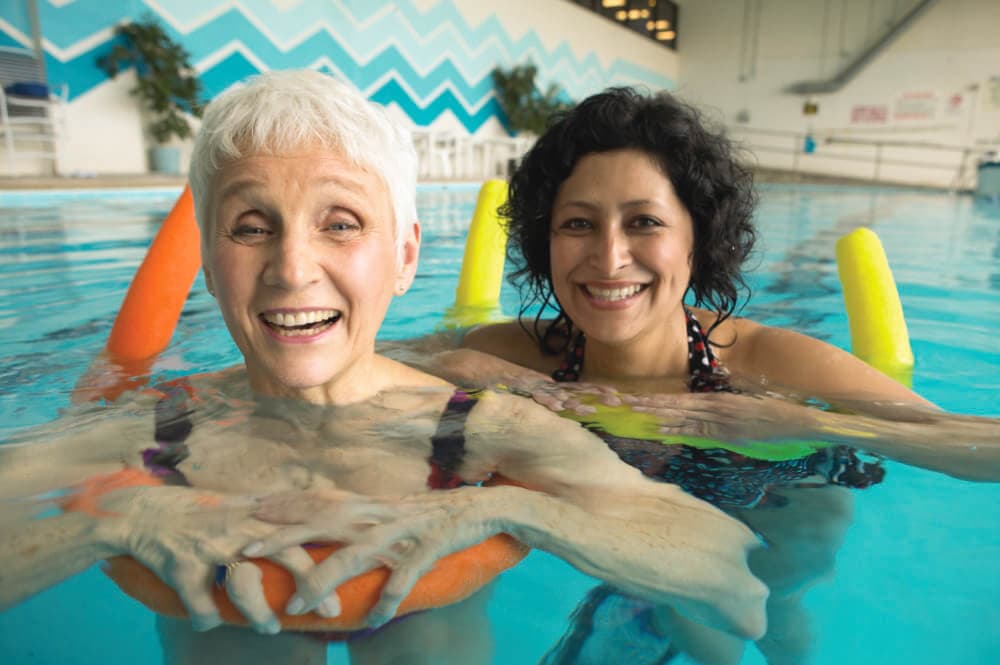 Two smiling women in a pool with pool noodles, one with short white hair, the other with short curly dark hair. - Home Instead