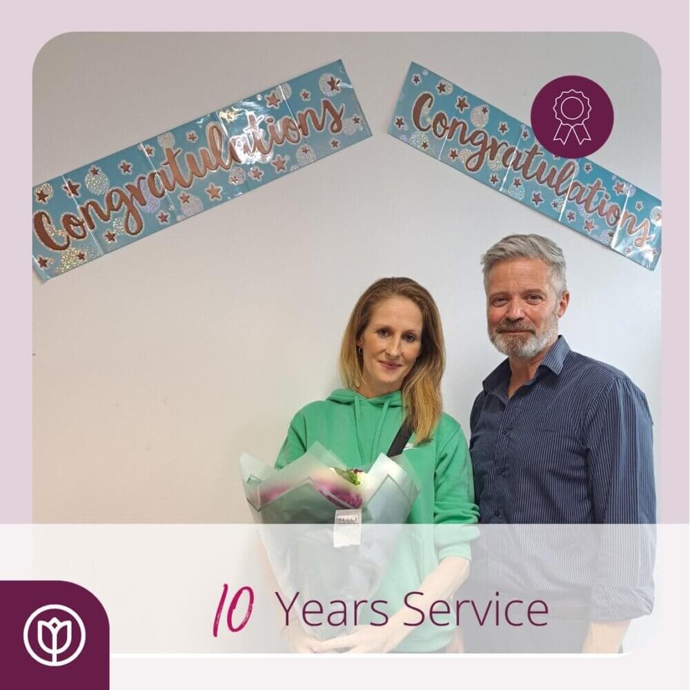 Two people stand together; one holds flowers. Banners reading "Congratulations" are in the background. Text says "10 Years Service. - Home Instead