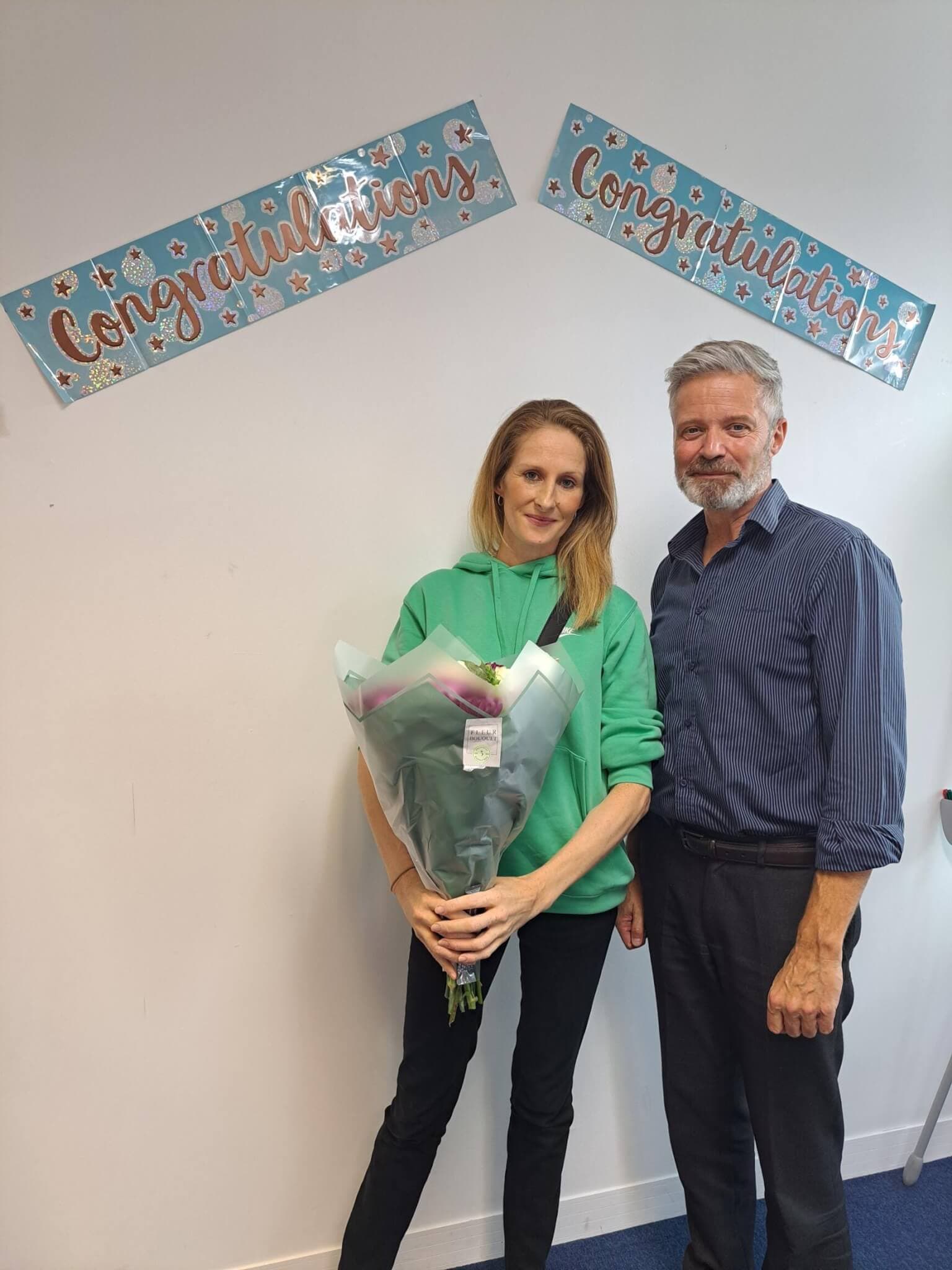 A woman holding a bouquet stands beside a man, with "Congratulations" banners on the wall behind them. - Home Instead