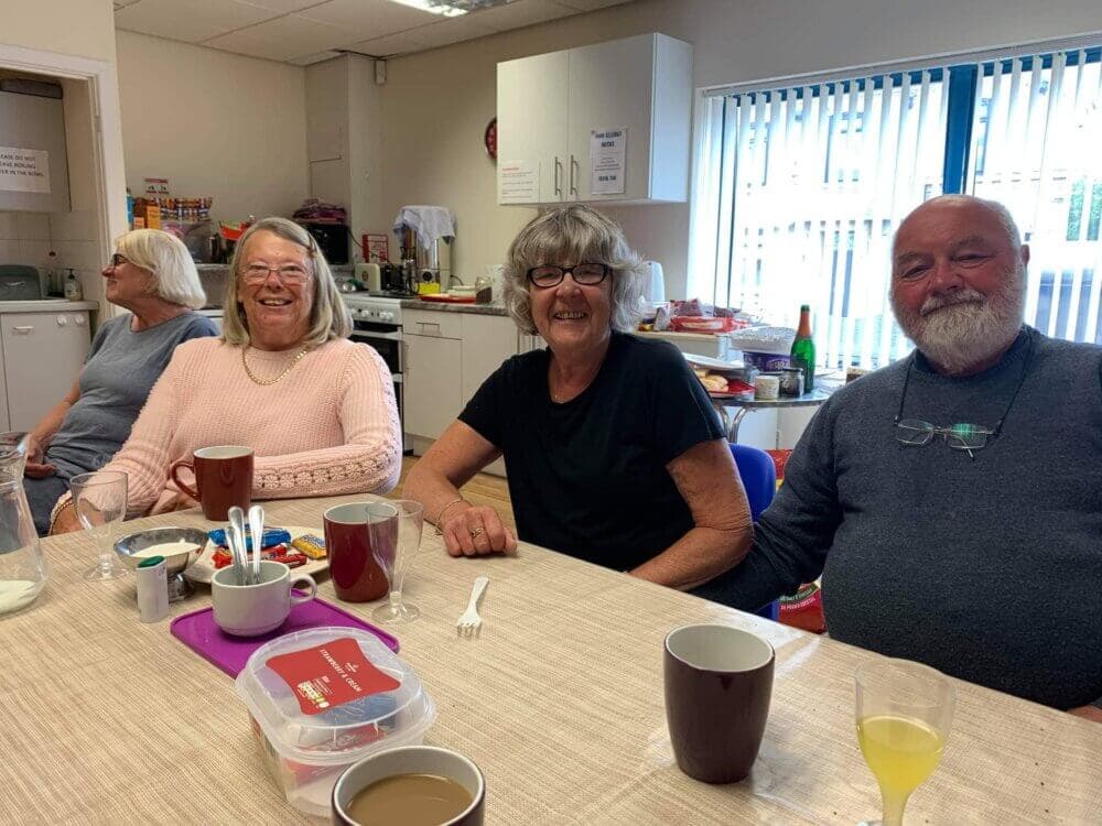 Four smiling people sitting around a table with drinks and snacks in a well-lit room. - Home Instead