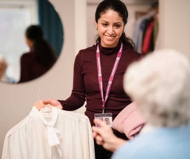 A woman smiles while holding a white blouse on a hanger, interacting with an older person in a blue shirt. - Home Instead