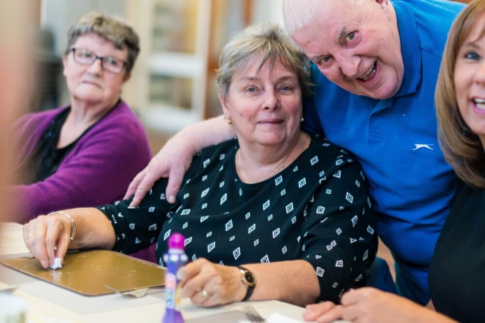 Three older adults sitting at a table, one man standing behind them with his arm around a woman, all smiling warmly. - Home Instead